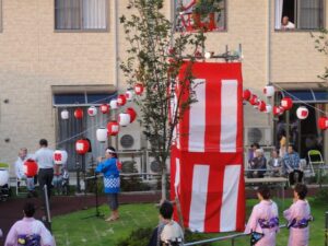 施設の庭で行われる夏祭りやイベント。入居者が大きな赤白の装飾オブジェの周りで楽しむ様子。